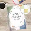 ADHD Task Hub daily planner on a wooden desk with a coffee cup, showing the calming abstract cover design.
