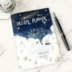 Dream Planner (1) The Dream Planner with a whimsical celestial cover design, resting on a marble desk next to a fountain pen.