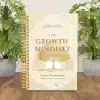The Growth Mindset Workbook displayed on a wooden desk between two green monstera plants, showcasing its elegant cover.